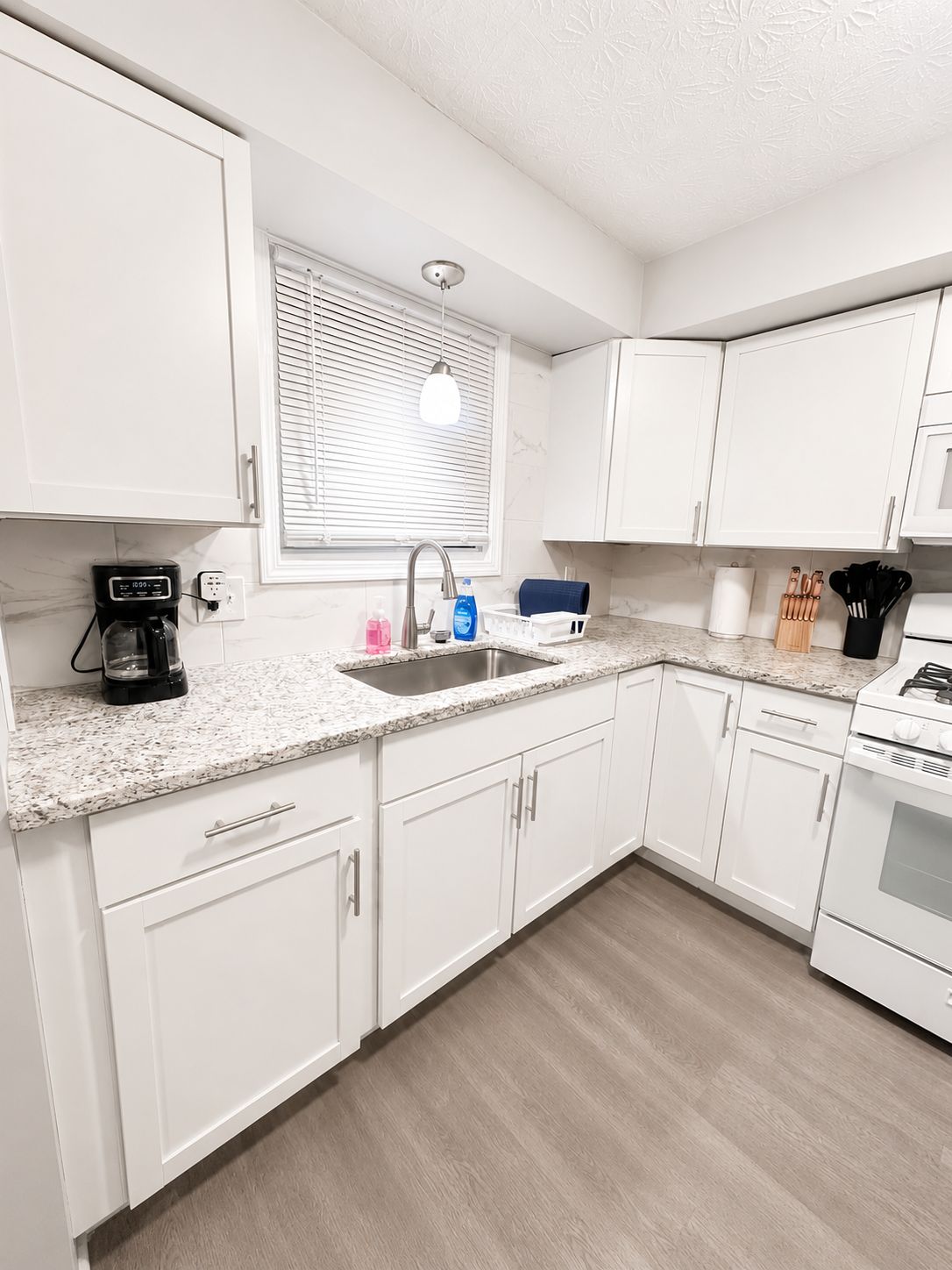A modern kitchen with white cabinets, a granite countertop, a built-in oven, and a window with blinds.