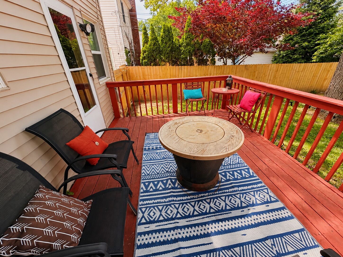 A wooden deck with a round table and chairs, a patterned rug, and a red railing.