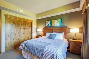 Un dormitorio con un amplio armario de madera, una cama con cubrecama a rayas, dos mesitas de noche con lámparas y una obra de arte colgada en la pared.
