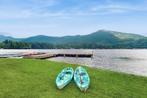 Eine grüne Wiese mit zwei Kajaks und einem Steg, der in einen See führt, mit Bergen im Hintergrund.