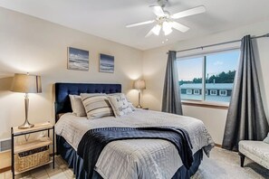 Ein Schlafzimmer mit Bett, Nachttischlampen, Deckenventilator, Blick auf Gebäude und einem Fenster mit Vorhängen.
