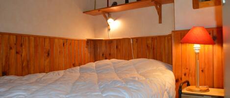A bedroom with a wooden headboard, a bedside table with a lamp, and a shelf with decorative items.