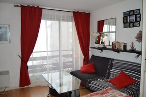 Una sala de estar con un sofá, una mesa de centro de vidrio, cortinas rojas, una ventana y una pared con cuadros.