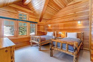 Een slaapkamer in een blokhut met twee houten bedden, een bureau en een raam met uitzicht op bomen.