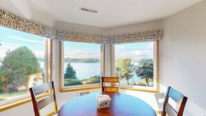 Comedor con una mesa redonda y sillas, ventanas amplias con vista a un lago y árboles, y un respiradero en el techo.