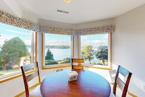 Une salle à manger avec une table ronde et des chaises, de grandes fenêtres donnant sur un lac et des arbres, et une bouche d’aération au plafond.