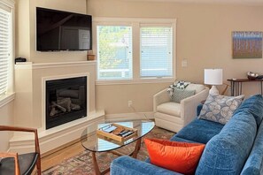 Ein Wohnzimmer mit Kamin, einem darüber montierten Fernseher, einem Sofa, einem Couchtisch und einem Fenster mit Jalousien.