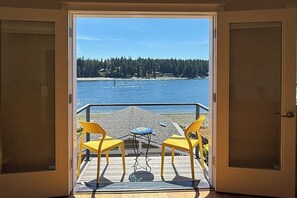 An open door leading to a balcony with two yellow chairs and a small table, overlooking a body of water and a forested shoreline. - Centrally located 2BR Oceanview - Fireplace (Suquamish)