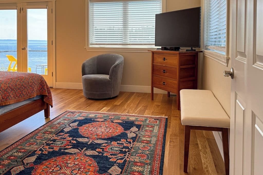 A bedroom with a bed, a chair, a television, a dresser, and a view of the ocean.