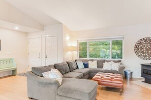 Ein Wohnzimmer mit Ecksofa, einer Bank, einem Kamin und einem großen Fenster mit Blick auf Grünflächen.