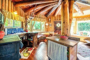 A rustic cabin interior with a wooden dining area, a kitchen with a sink, and a large window offering a view of the outdoors.
