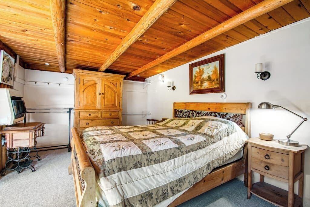 A bedroom with a wooden ceiling, a bed with a floral quilt, a wooden nightstand, and a wardrobe.