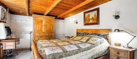 A bedroom with a wooden ceiling, a bed with a floral quilt, a wooden nightstand, and a wardrobe.