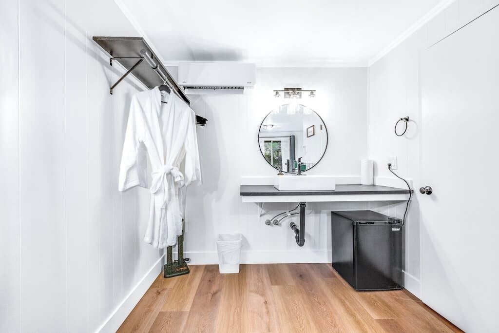 A modern bathroom with a white vanity, a round mirror, a towel rack, and a small black trash bin.