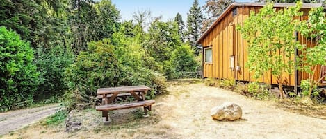 A wooden cabin with a picnic table and a dirt path leading to it, surrounded by dense forest.