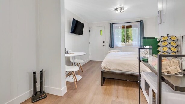 A compact room with a bed, a small table, a chair, a TV mounted on the wall, and a window with curtains.