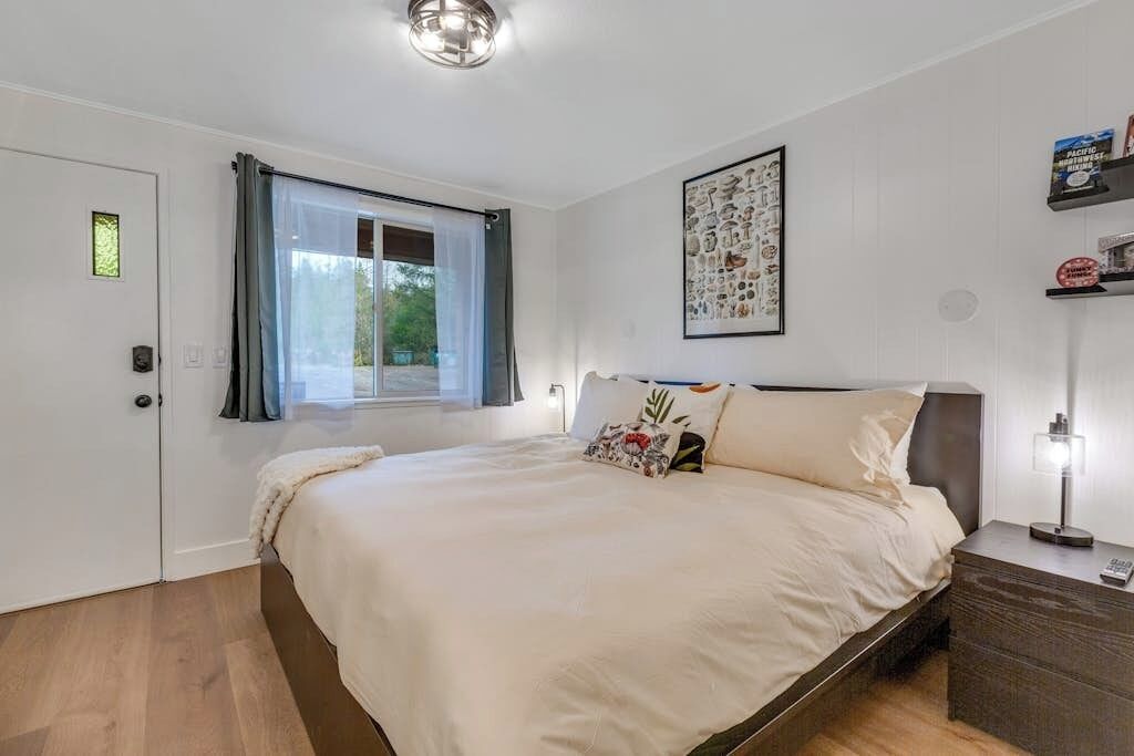 A bedroom with a bed, a window with curtains, a bedside table with a lamp, and a wall with a framed picture.