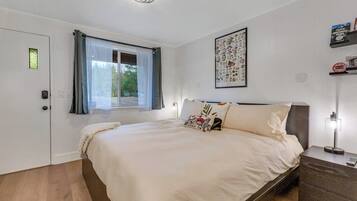 A bedroom with a bed, a window with curtains, a bedside table with a lamp, and a wall with a framed picture.