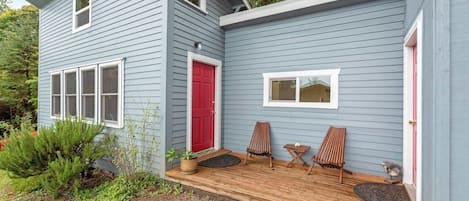 Uma casa de dois andares com um deck de madeira, uma porta vermelha e duas cadeiras.