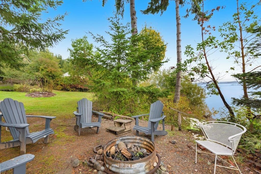 Een zitgedeelte aan het meer met Adirondack-stoelen en een metalen stoel, omgeven door bomen en een vuurkuil.