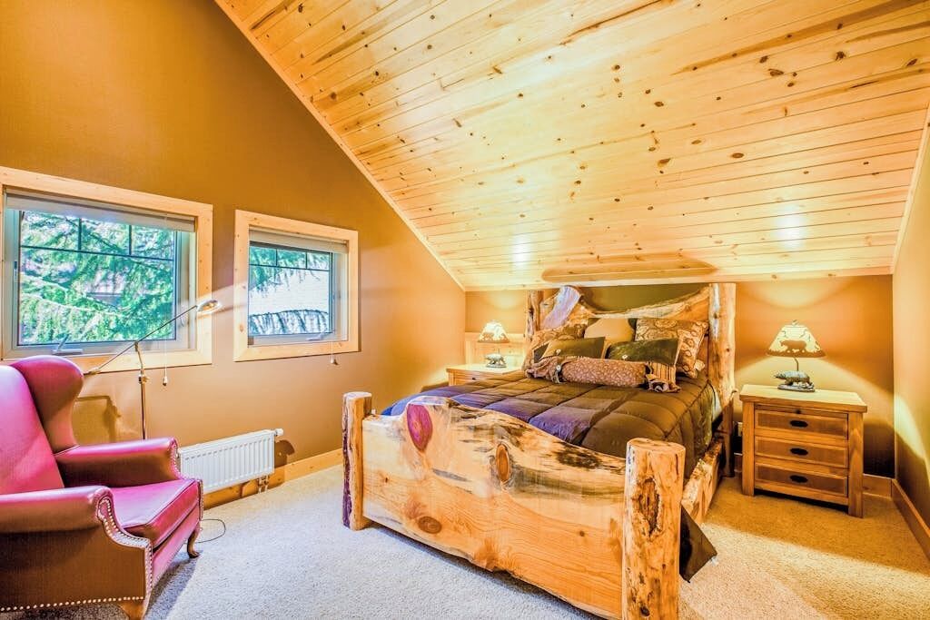 A bedroom with a sloped ceiling, a wooden bed frame, a red chair, two bedside tables with lamps, and a window with a view of greenery.