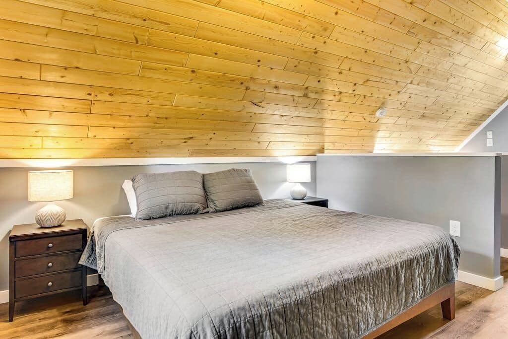 A bedroom with a sloped ceiling, a bed with a grey bedspread, two bedside lamps, and a nightstand with drawers.