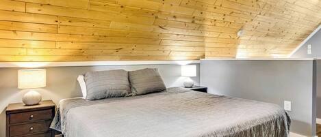 A bedroom with a sloped ceiling, a bed with a grey bedspread, two bedside lamps, and a nightstand with drawers.