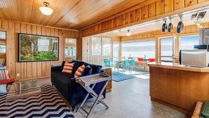 A wooden interior with a sofa, a dining area, and a view through large windows.