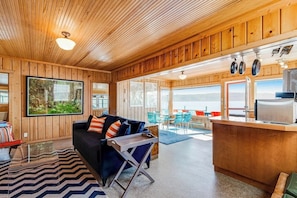 A wooden interior with a sofa, a dining area, and a view through large windows.