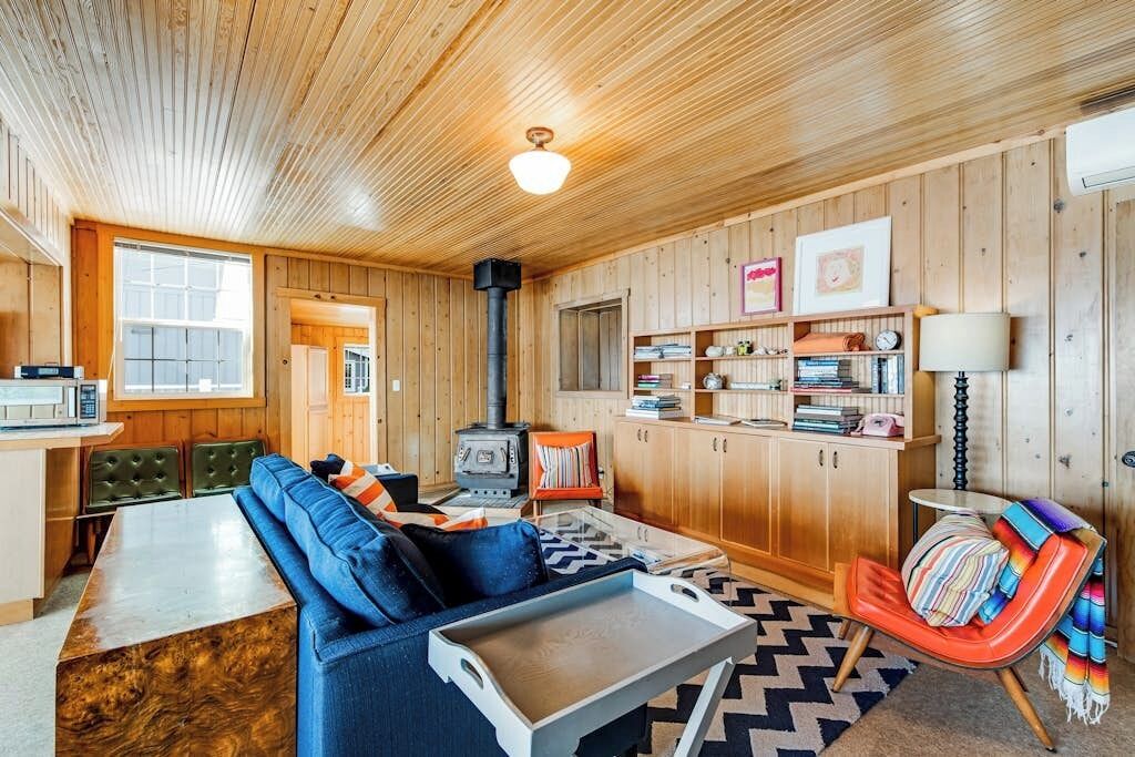 A cozy cabin interior with a wooden ceiling, a sofa, a dining table, a kitchen area, and a fireplace.