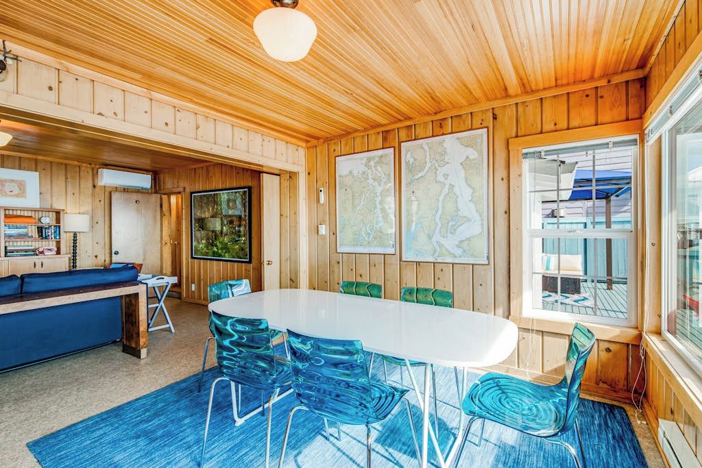 A wooden cabin interior with a dining table, chairs, a sofa, and a large window.