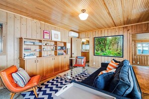 A wooden-paneled room with a sofa, a chair, a bookshelf, and a large window with a view of greenery.