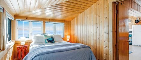 Un dormitorio de cabaña de madera con una cama, ventanas y una puerta que da a una cocina.