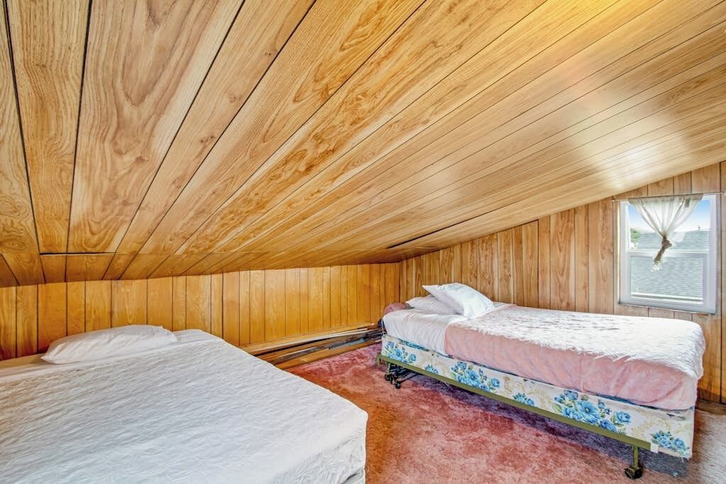 A room with wooden walls and ceiling, two beds with floral bedding, and a window with a view.