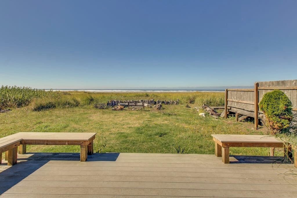 A wooden deck with two benches overlooking a grassy area with a wooden fence and a view of the beach.