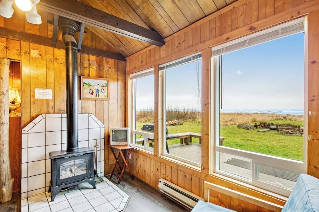 A wooden cabin interior with a fireplace, a television, and large windows overlooking a grassy area.