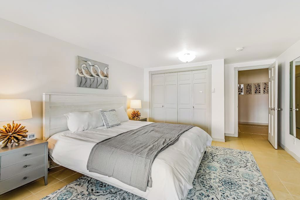 A bedroom with a bed, bedside tables, a nightstand, a wall-mounted artwork, a wardrobe, and a patterned rug.