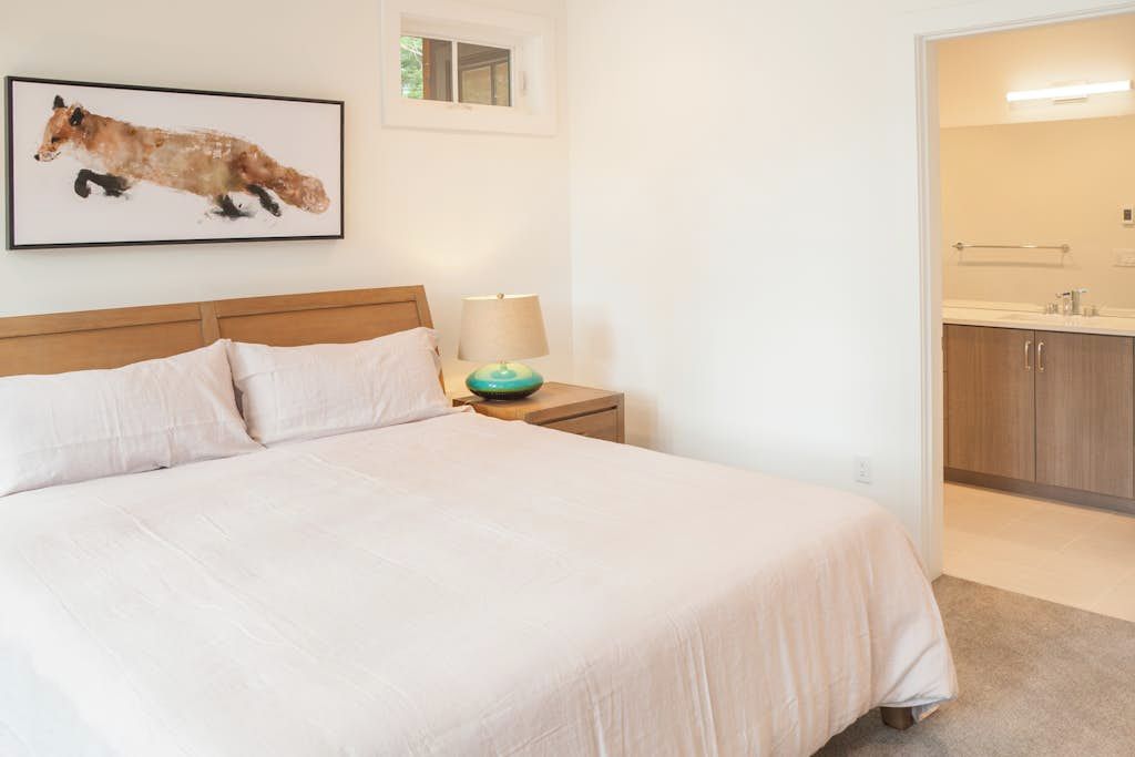 A bedroom with a bed, a nightstand, a lamp, a framed picture, and a bathroom with a sink and mirror.