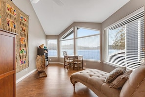 A room with a wooden floor, a cushioned seating area, a dining table with chairs, and large windows with blinds. - 2BR lakefront home - kayaks, grill, beach access (Olympia)