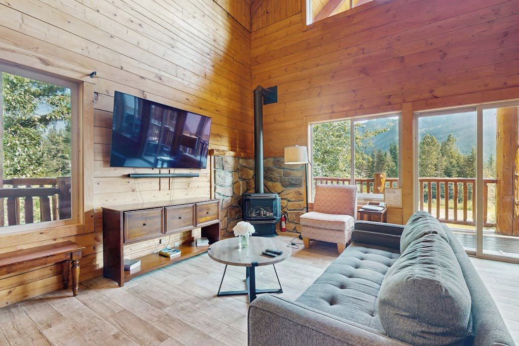 A cozy cabin interior with a television, fireplace, sofa, and large windows offering a view of the outdoors.