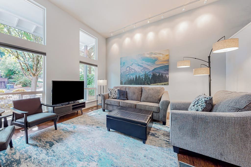 A modern living room with a large flat-screen TV, a grey sofa set, a coffee table, and a large window providing a view of greenery.