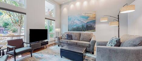 A modern living room with a large flat-screen TV, a grey sofa set, a coffee table, and a large window providing a view of greenery.