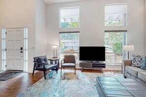 A modern living room with a flat-screen TV, two armchairs, a sofa, and a large window providing natural light.