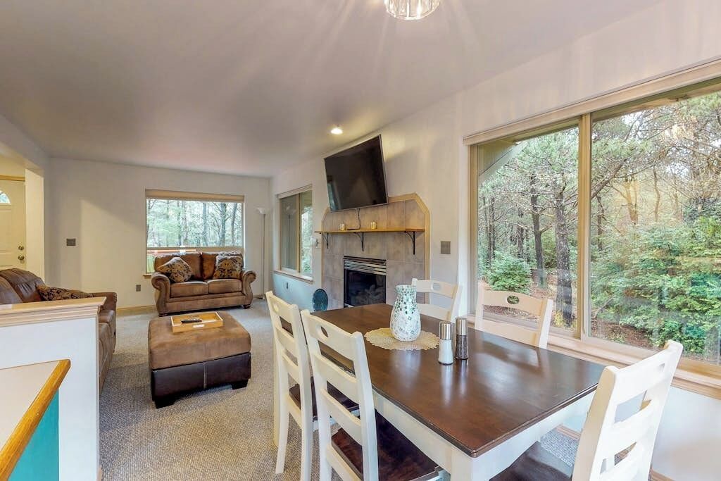 Un salon moderne doté d’une cheminée, d’un coin repas et d’une grande fenêtre donnant sur la forêt.