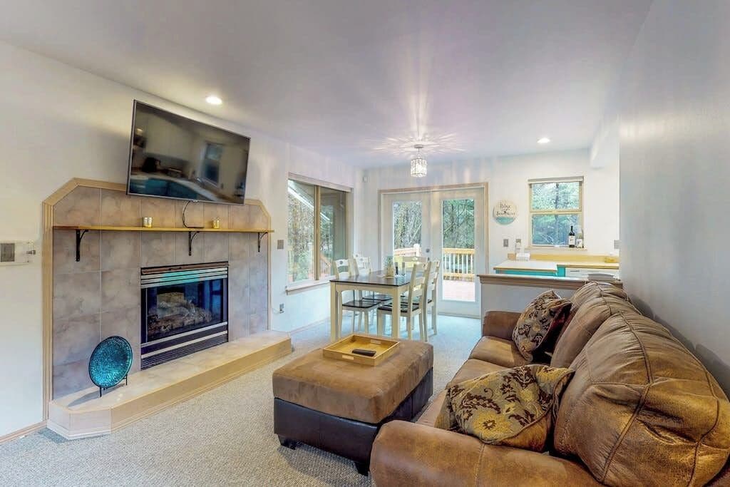 A modern living room with a fireplace, a flat-screen TV mounted above it, a brown leather sofa, and a dining area with a glass table and chairs.