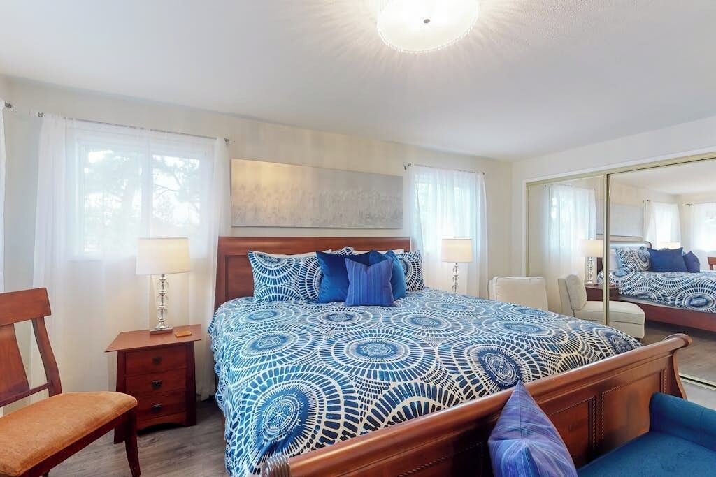 Une chambre d’hôtel avec un grand lit, une tête de lit en bois, une table de chevet, une chaise, un miroir et une fenêtre avec des rideaux.