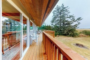 A wooden deck with glass railings, outdoor seating, and a view of a coniferous tree and open field. - Spacious 3BR Bayview Dog Friendly - Balcony (Westport)