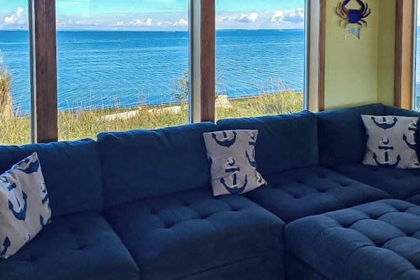 A room with a large window overlooking a body of water, a dark blue sofa with matching cushions, and a wall-mounted decorative item. - 3BR beachfront paradise with outstanding views (Sequim)