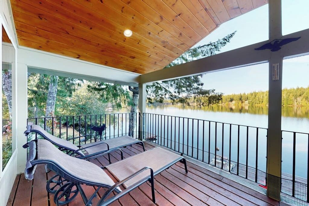 A covered outdoor deck with lounge chairs, a railing, and a view of a lake.