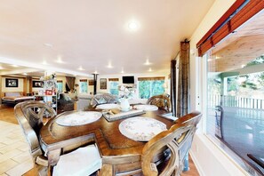 Un coin repas avec une table et des chaises en bois, un téléviseur fixé au mur et une vue sur l’extérieur grâce à de grandes fenêtres.